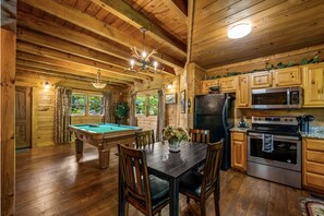 A wooden cabin interior with a pool table, dining table, and kitchen area including a refrigerator and stove.