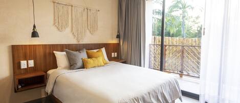 Un dormitorio con cabecera de madera, cama con ropa de cama blanca, mesita de noche, aire acondicionado de pared y vistas a una terraza de madera y palmeras a través de un ventanal grande.