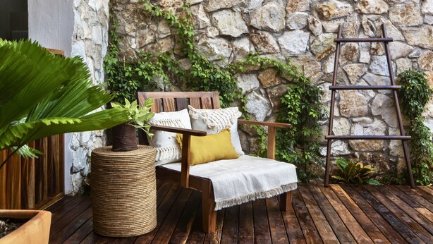 Eine Holzterrasse mit einer Steinmauer, einem Holzstuhl mit Kissen, einem gewebten Hocker und einer Metallleiter.