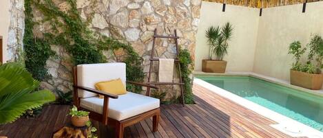 Terraza de madera con un muro de piedra, una silla de descanso, una piscina pequeña y plantas en macetas.