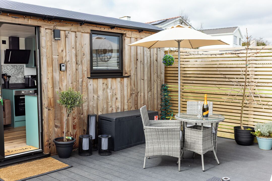 Une terrasse en bois avec des meubles d’extérieur, une porte vitrée donnant sur une cuisine et une clôture en bois.