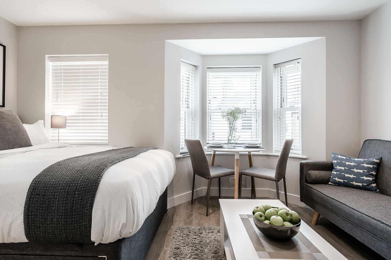 A modern bedroom with a large bed, a desk with chairs, a sofa, and a view through a window.