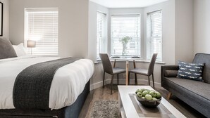 A modern bedroom with a large bed, a desk with chairs, a sofa, and a view through a window. - Central Cambridge Studios - Tailored Stays (Cambridge)
