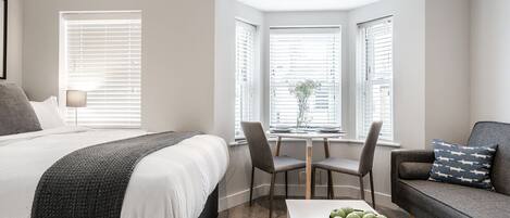 A modern bedroom with a large bed, a desk with chairs, a sofa, and a view through a window.