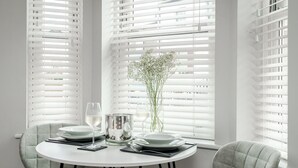 A dining area with a round table set for two, white blinds on the windows, and a vase with flowers on the table. - Central Cambridge Studios - Tailored Stays (Cambridge)