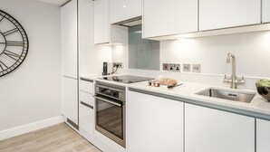A modern kitchen with white cabinetry, a built-in oven, a stainless steel sink, and a large wall clock. - Central Cambridge Studios - Tailored Stays (Cambridge)