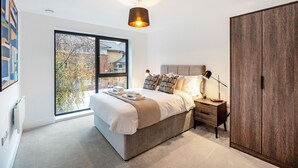 A modern bedroom with a large window, a bed with a headboard, a bedside table, and a wardrobe. - Lacon House 2-bedroom apartment (Cambridge)