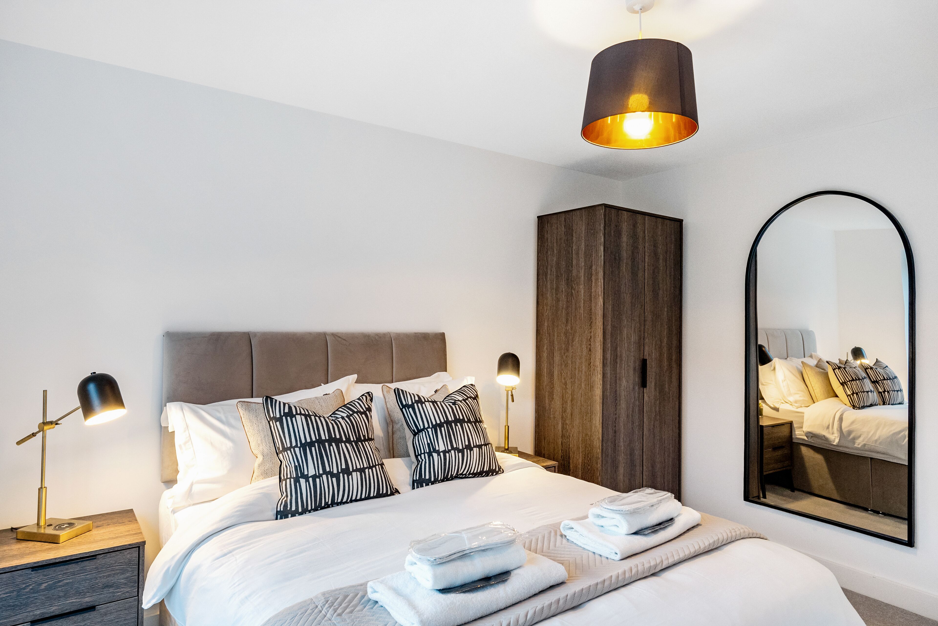 Un dormitorio moderno con una cama grande, un armario, una mesita de noche con lámpara y un espejo en la pared.