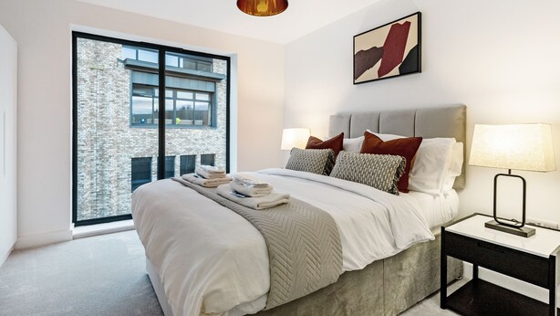 A modern bedroom with a large bed, a nightstand, a lamp, and a window with a view of a brick building. - Lacon House 1-bedroom apartment (Cambridge)