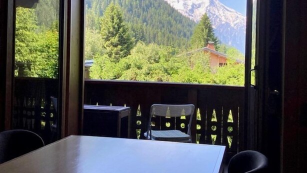 Ein Zimmer mit Tisch und Stühlen, ein Fenster mit Blick auf einen Berg und Wald.