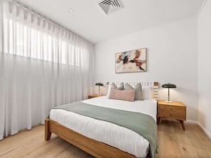 A modern bedroom with a wooden bed, white bedding, a green throw, two bedside lamps, and a painting on the wall.