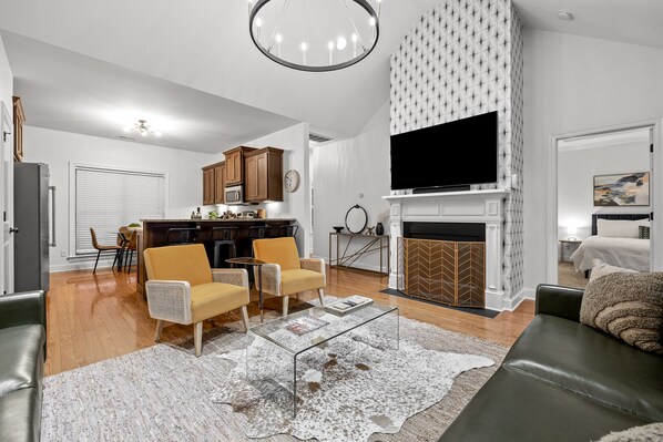 A modern living room with a fireplace, a television, and a dining area with wooden cabinets and a kitchen in the background.