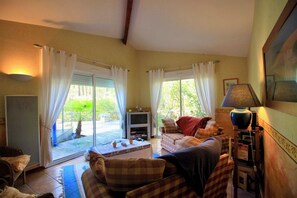 A living room with a sofa, a television, and a table with books. There are curtains on the windows, and a lamp is on a side table. - House with pool and terrace facing the Golf G0275 (Lacanau-Ocean)