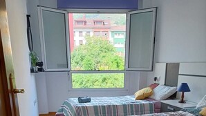 Une chambre avec deux lits, une fenêtre donnant sur des bâtiments et de la verdure, et une table de chevet avec une lampe.