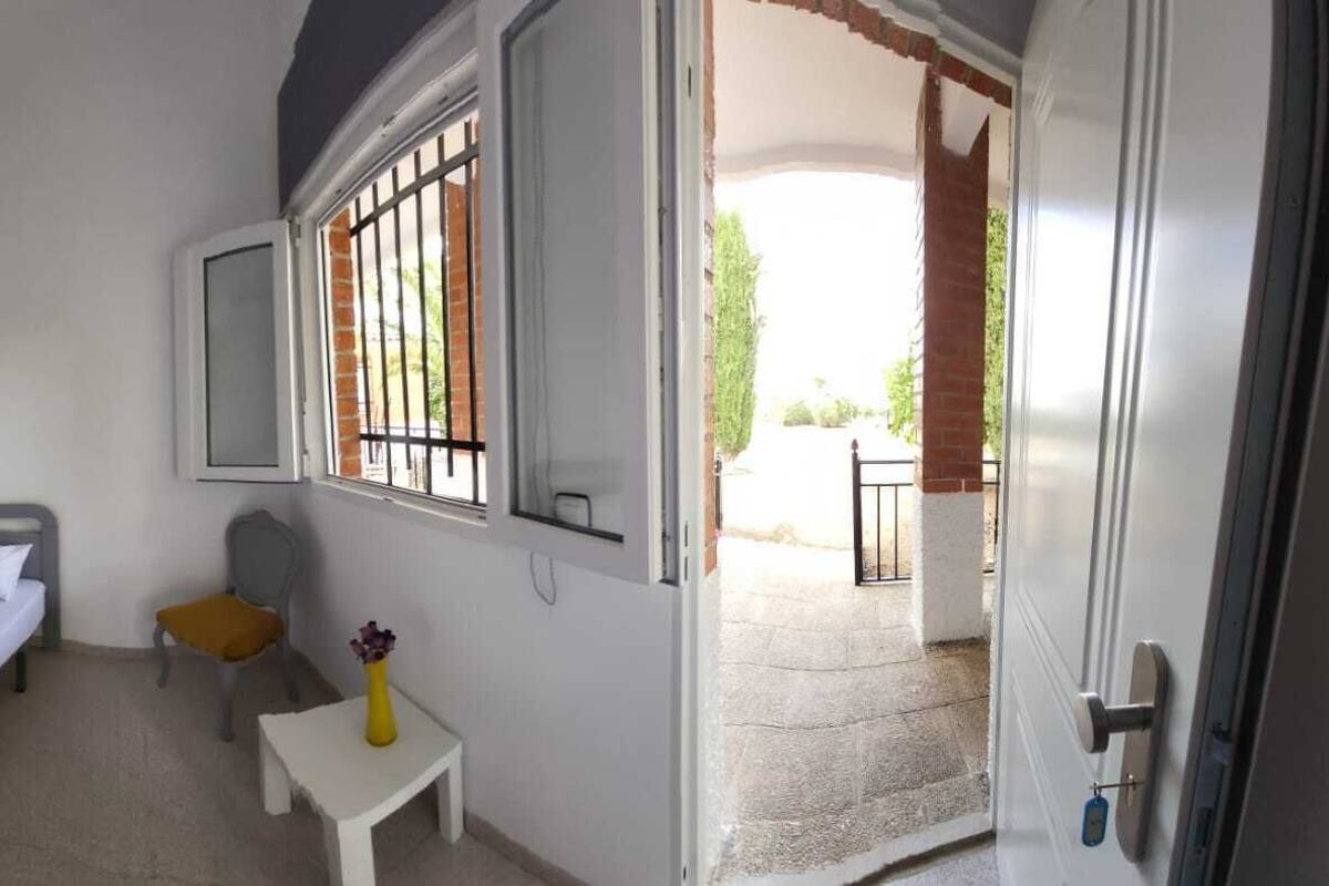 Ein Hotelzimmer mit zwei Einzelbetten, einem kleinen Tisch mit einer Vase, einem Stuhl, einem Fenster mit Gitterstäben und einer zur Außenwelt führenden Tür.