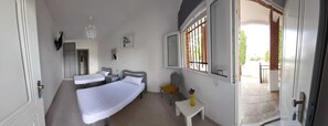 Une chambre d’hôtel avec deux lits simples, une petite table avec un vase, une chaise, une fenêtre avec des barreaux et une porte donnant sur l’extérieur.