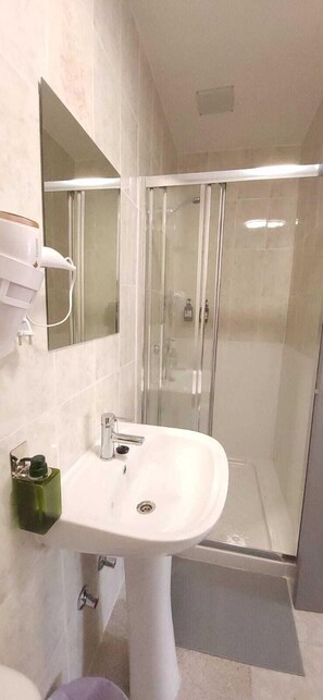 Une salle de bain avec un lavabo sur colonne blanc, un miroir, une cabine de douche en verre et un distributeur de savon vert.