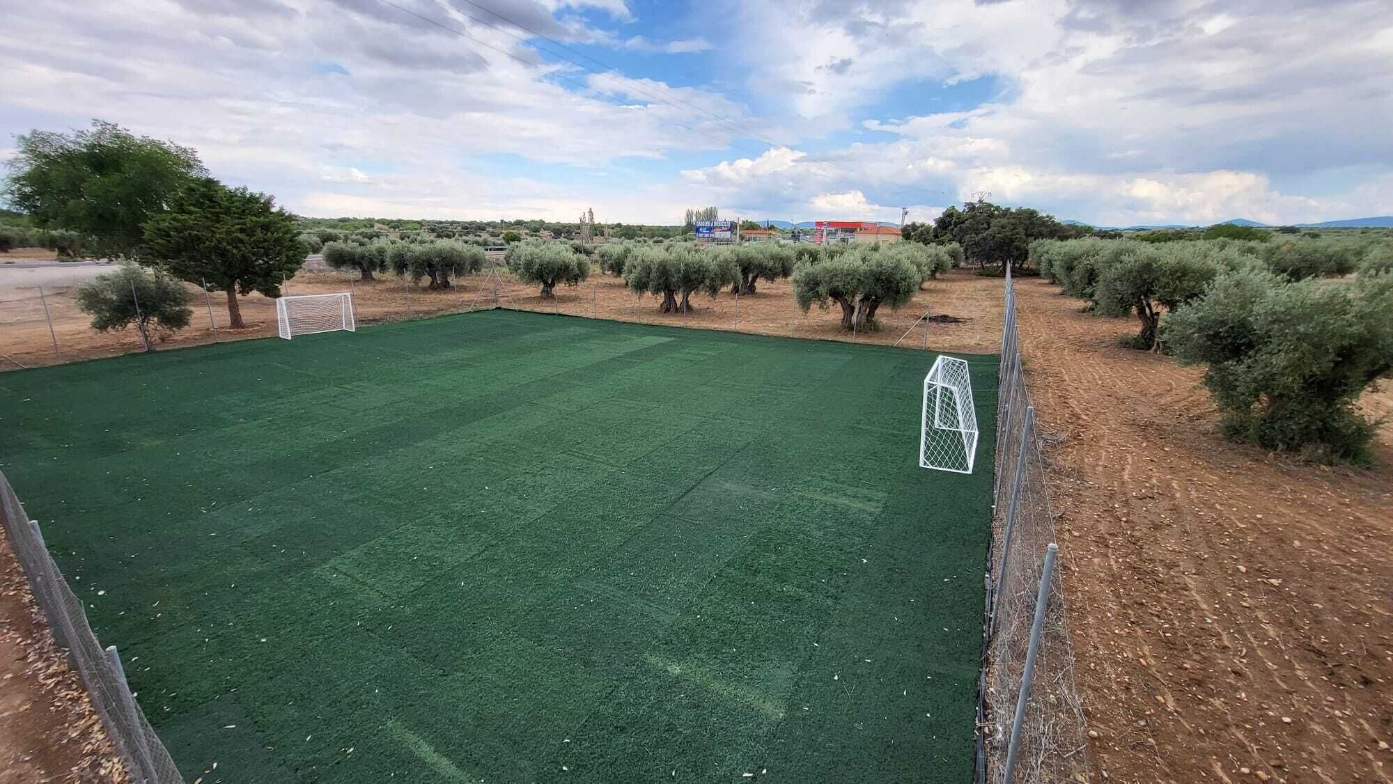 Un terrain de football clôturé, avec des buts, entouré d’arbres et avec un bâtiment visible au loin.