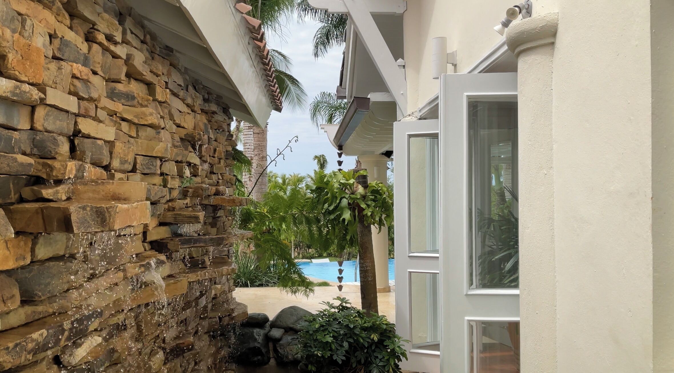 A stone wall with a water feature, a pool, and a view of a palm tree.