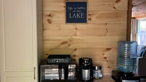 Une cuisine compacte avec un mur en bois, une machine à café, un distributeur d’eau et un panneau encadré sur lequel on peut lire : « La vie est plus belle au LAKE HOUSE ».