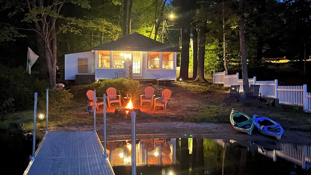 Une cabane avec une jetée s’avançant dans un lac paisible, entourée d’arbres et d’une clôture en bois peinte en blanc.