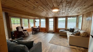 L’intérieur d’une cabane est doté d’un plafond en bois, d’un coin repas avec une table et des chaises, d’un canapé et offre une vue sur un lac grâce à de grandes fenêtres.