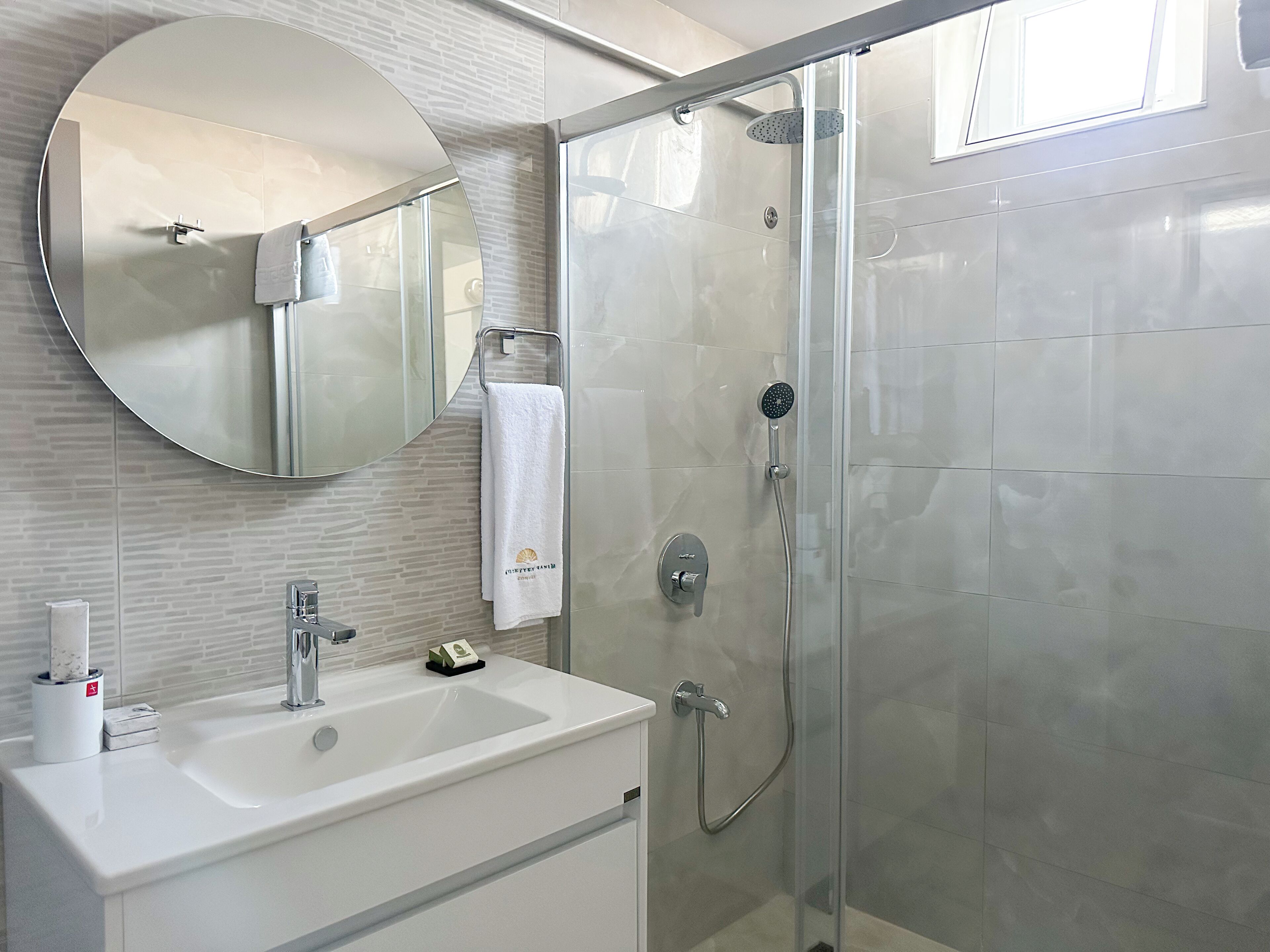 Un baño moderno con ducha de vidrio, lavamanos blanco y espejo redondo.