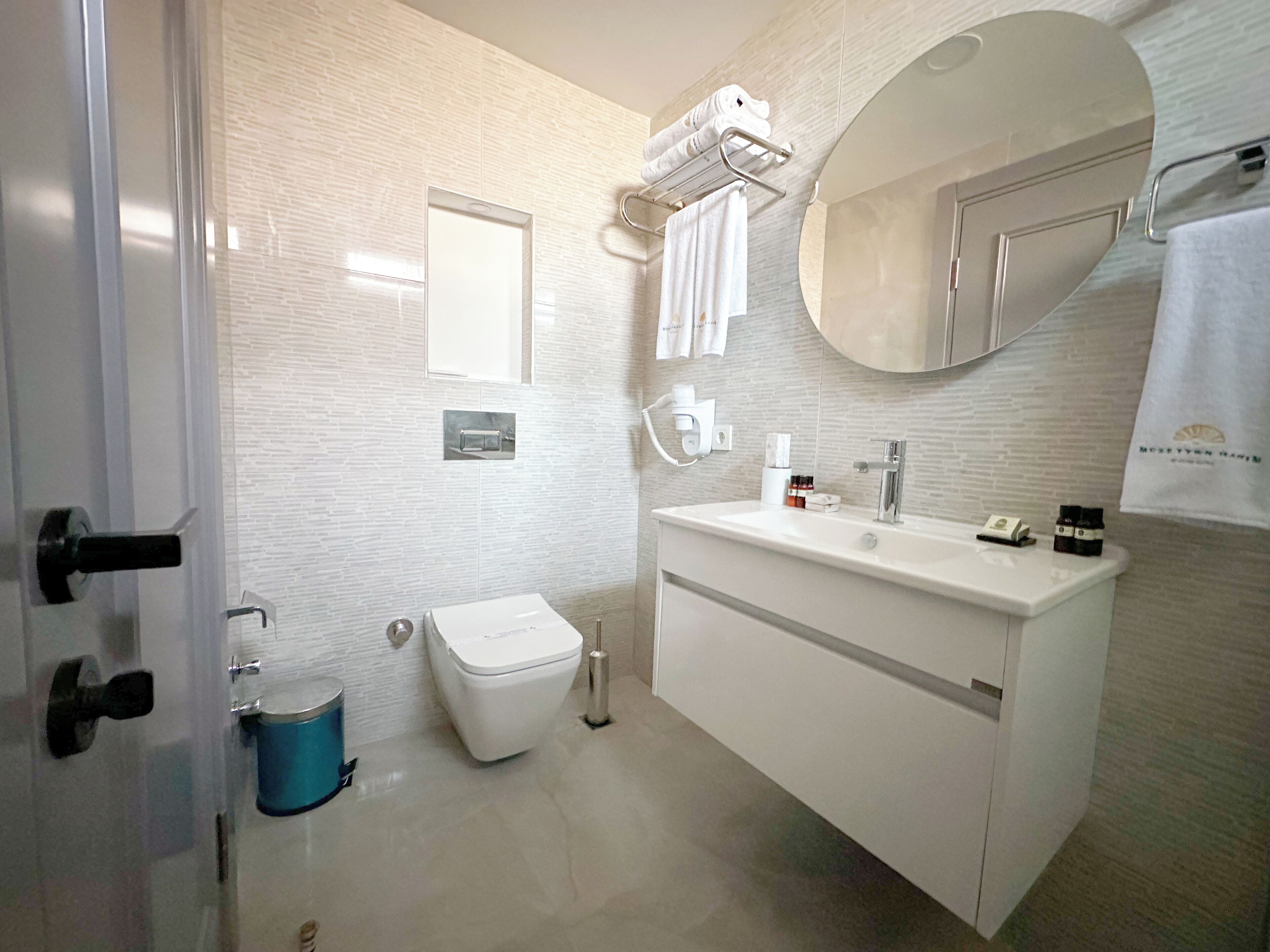 A modern bathroom with a white sink, mirror, and toilet. There is a towel rack above the sink and a towel hanging on it.