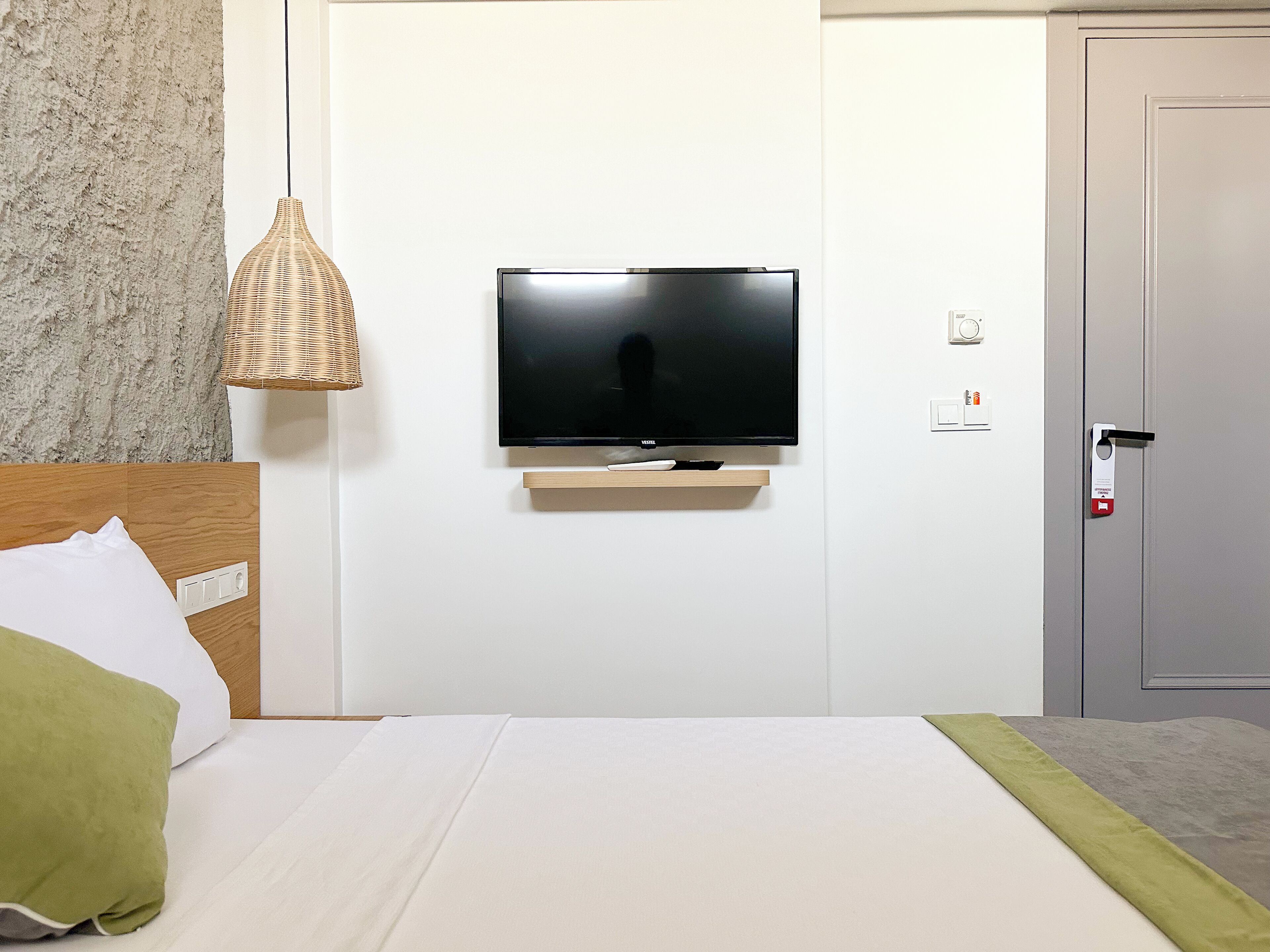 A hotel room with a flat-screen TV mounted on a wall, a wooden headboard, a green pillow, and a white bedspread.