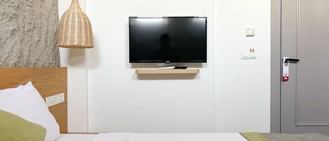A hotel room with a flat-screen TV mounted on a wall, a wooden headboard, a green pillow, and a white bedspread.