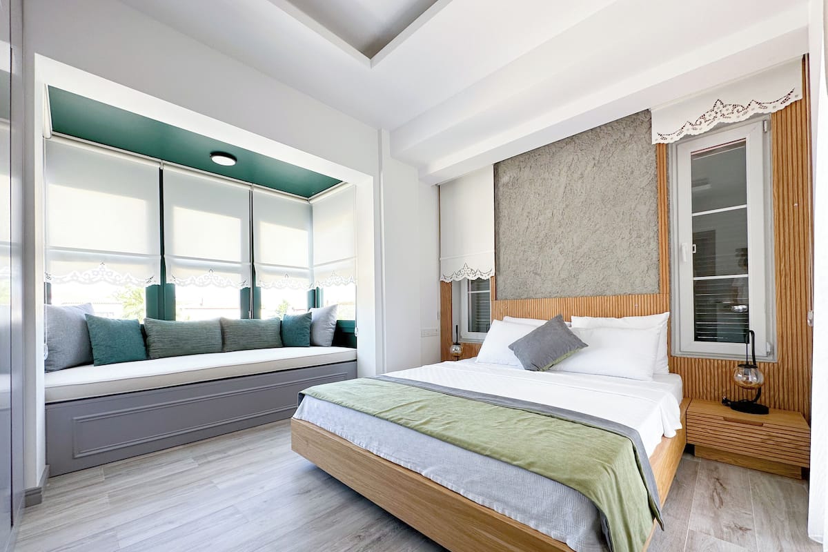 Une chambre avec un grand lit, un coin fenêtre agrémenté de coussins, une table de chevet et une lampe.