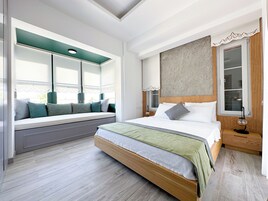 Un dormitorio con una cama grande, un rincón con ventanal con cojines, una mesita de noche y una lámpara.