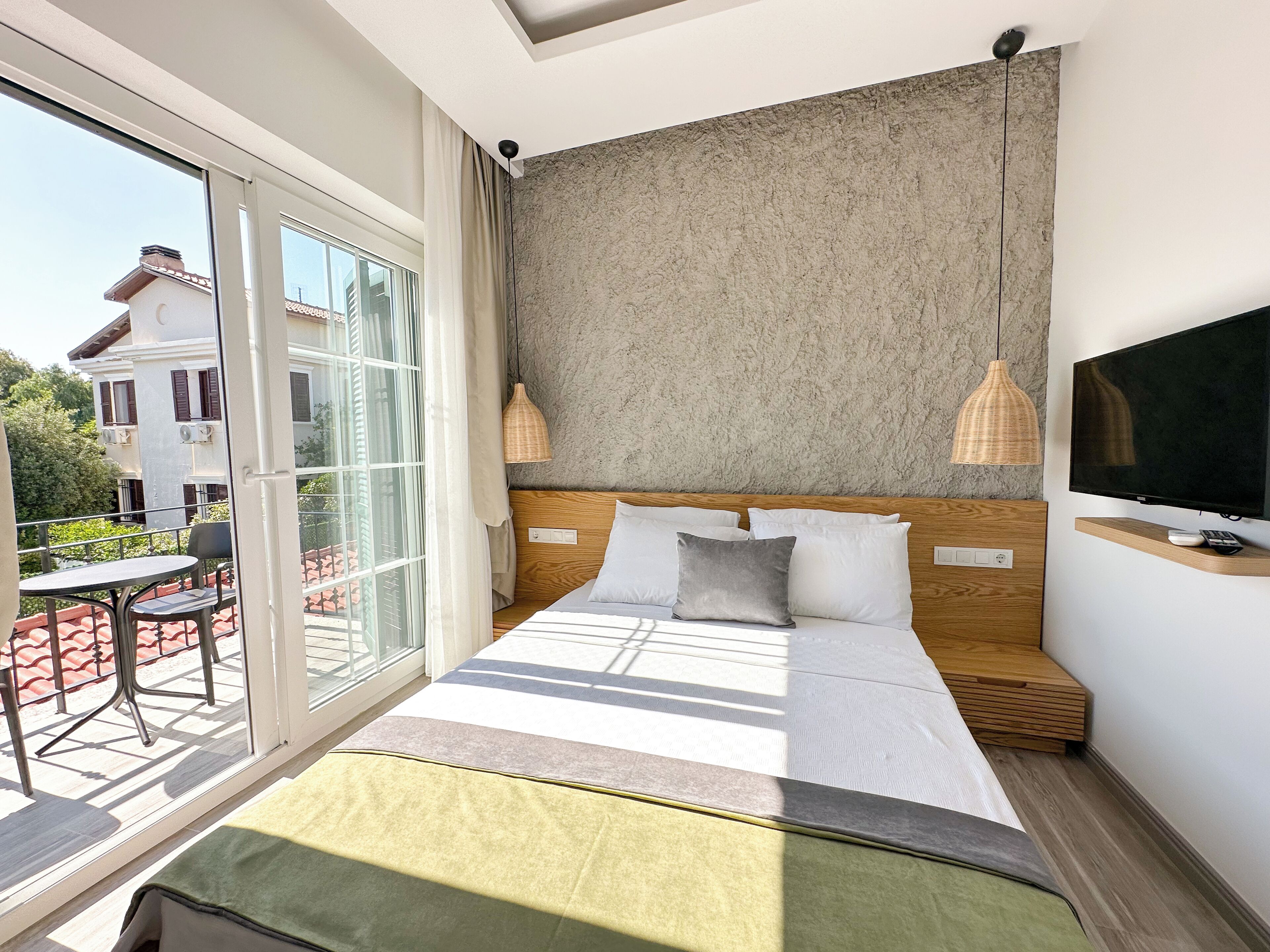 A modern hotel room with a large bed, a TV, and a balcony with outdoor seating.