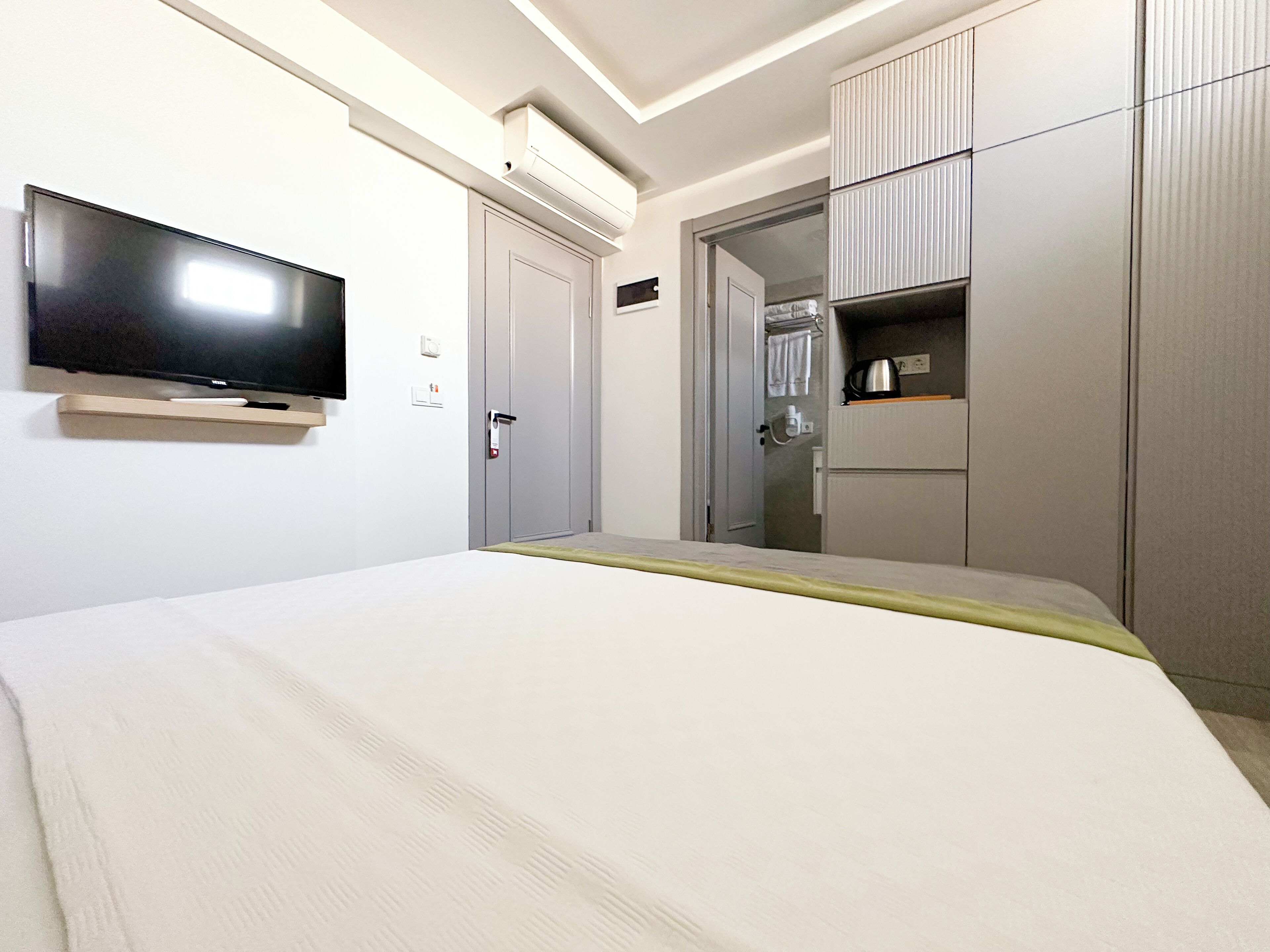 A hotel room with a bed, a flat-screen TV mounted on the wall, a wardrobe, and a small kitchenette area.