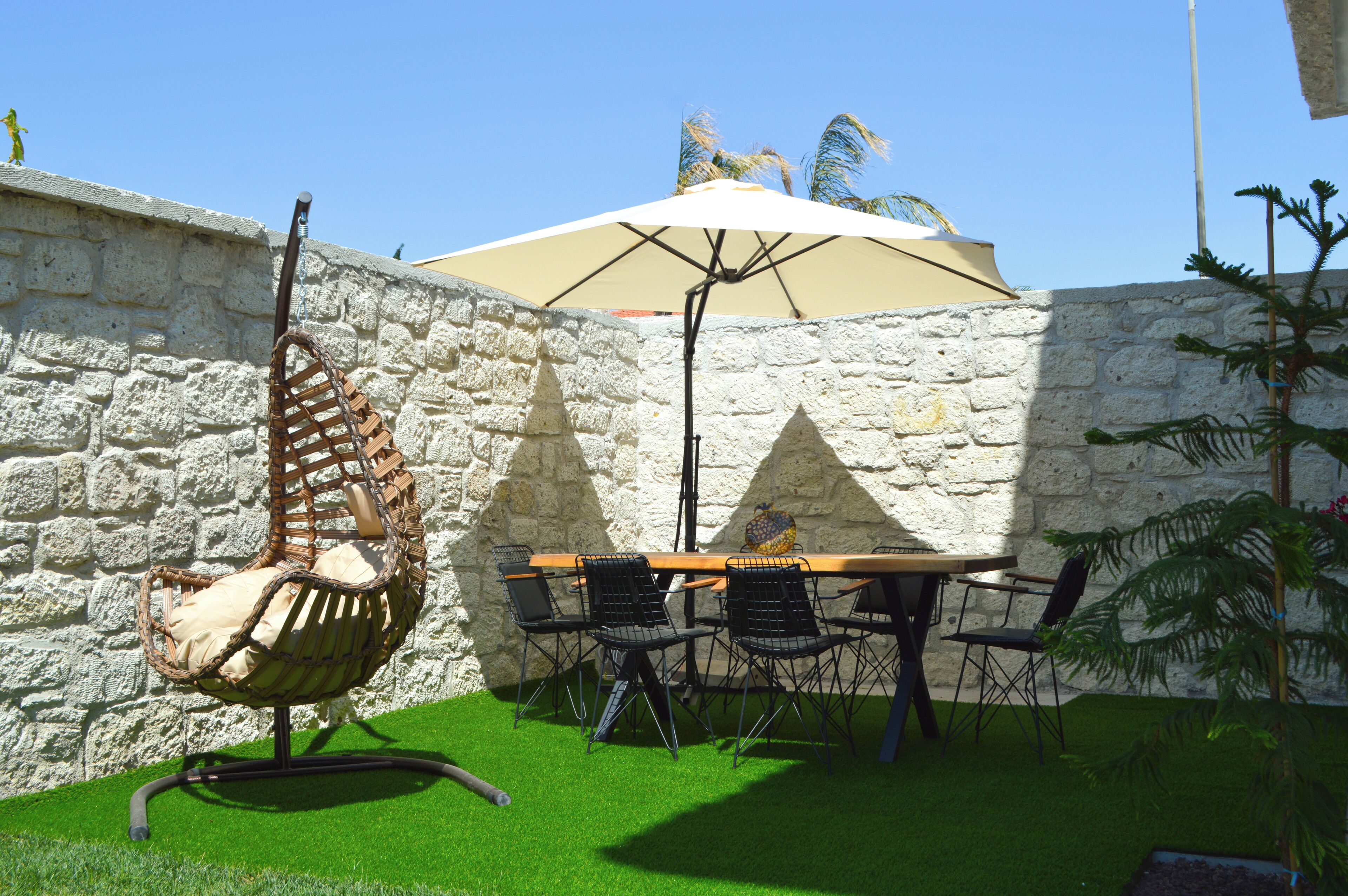 Un patio con un muro de piedra, una silla colgante, una mesa con sillas y una sombrilla.
