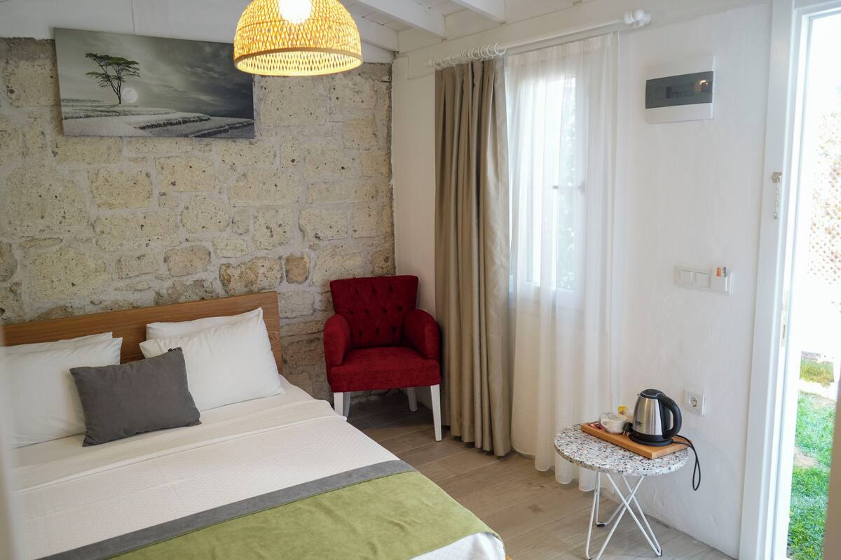 Une pièce avec un lit, une chaise rouge, une petite table avec une bouilloire et un tableau accroché au mur.