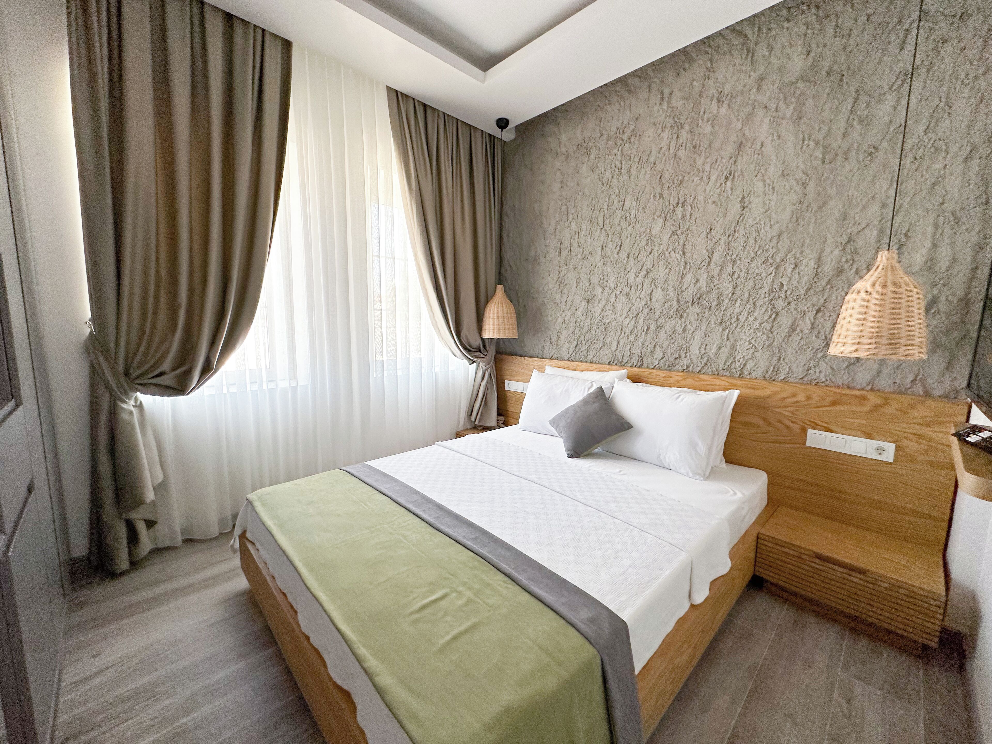A neatly made bed with a wooden headboard, a green throw blanket, and two pillows. There is a nightstand with a lamp and a power outlet. The room has a textured wall and a window with sheer curtains.