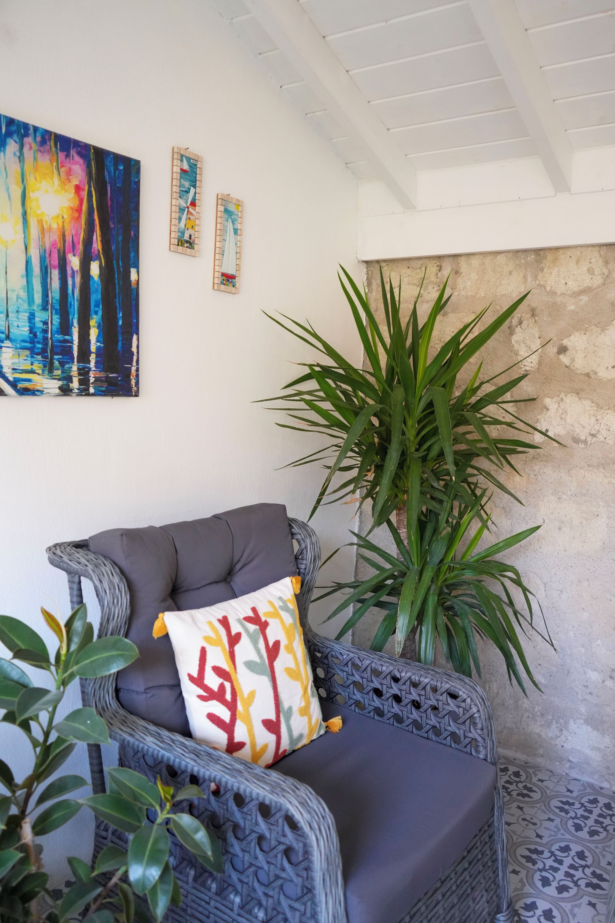 A wicker chair with a cushion, a large potted plant, and a colorful abstract painting on the wall.
