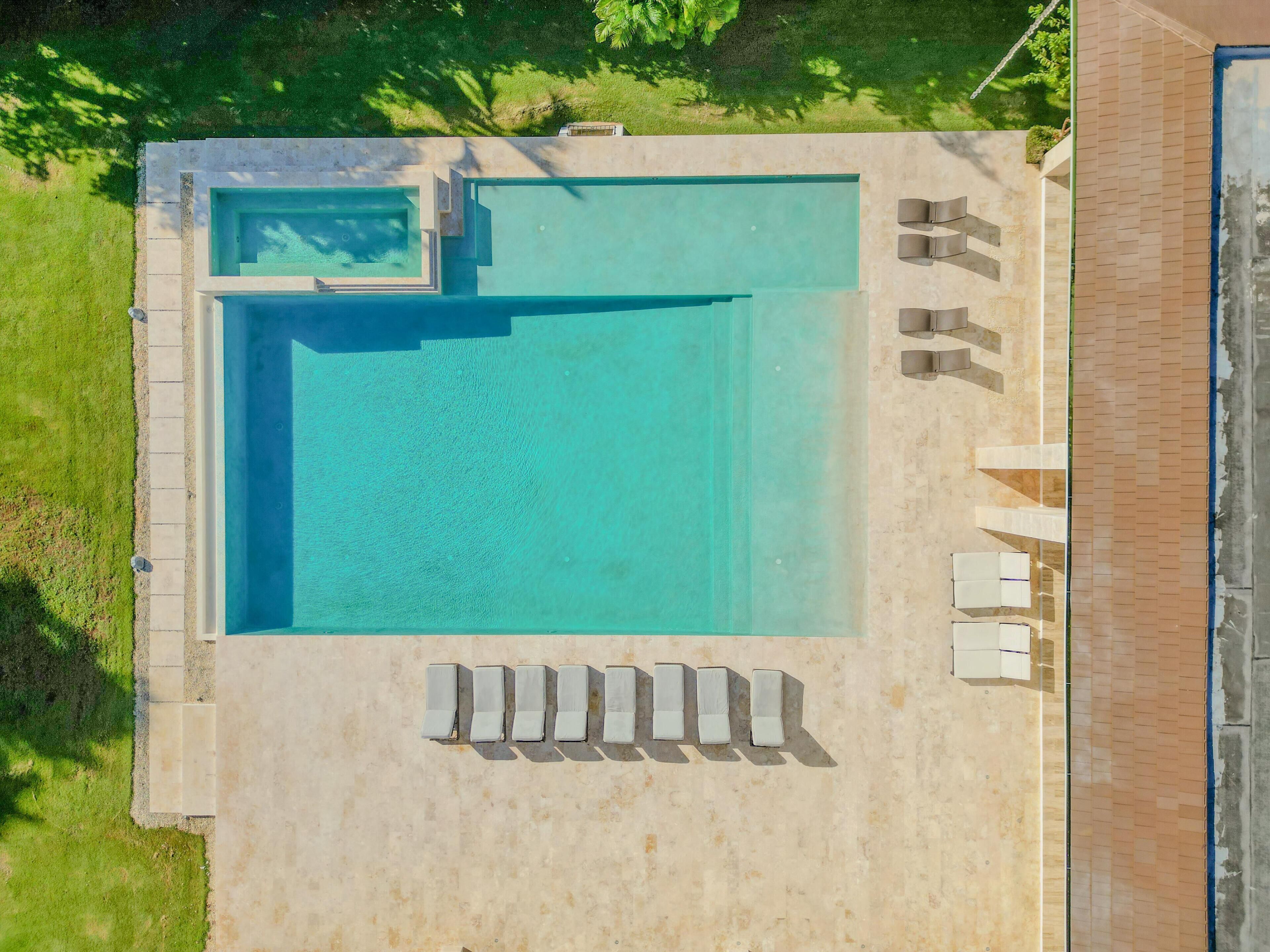 An aerial view of a rectangular swimming pool with a small adjacent section, surrounded by a light-colored deck. There are five lounge chairs aligned along the deck's edge.
