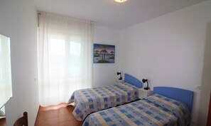 A bedroom with two beds, a window with curtains, a painting on the wall, and a desk with a chair.