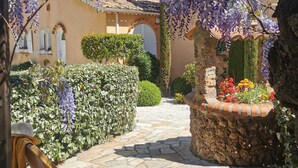 Un patio con una pérgola cubierta de flores moradas, un banco de piedra y una pequeña zona ajardinada con una jardinera de ladrillo.