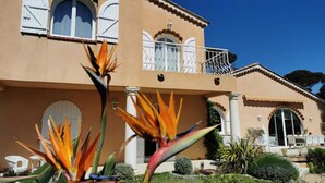 Un edificio de dos pisos con un balcón, ventanas arqueadas y un jardín bien cuidado con plantas tropicales.