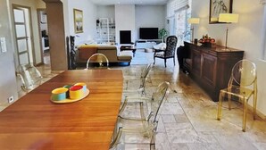 Un comedor amplio con una larga mesa de madera y sillas traslúcidas, una cocina al fondo, y una sala de estar con sofá y lámpara.
