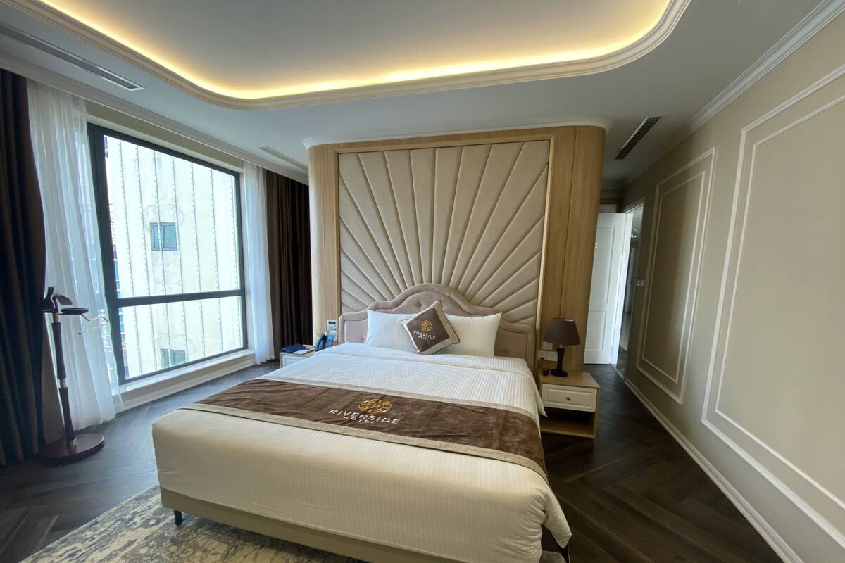 Una habitación de hotel con una cama grande, cabecera con diseño de abanico, mesita de noche, lámpara y vista a un edificio a través de una ventana.
