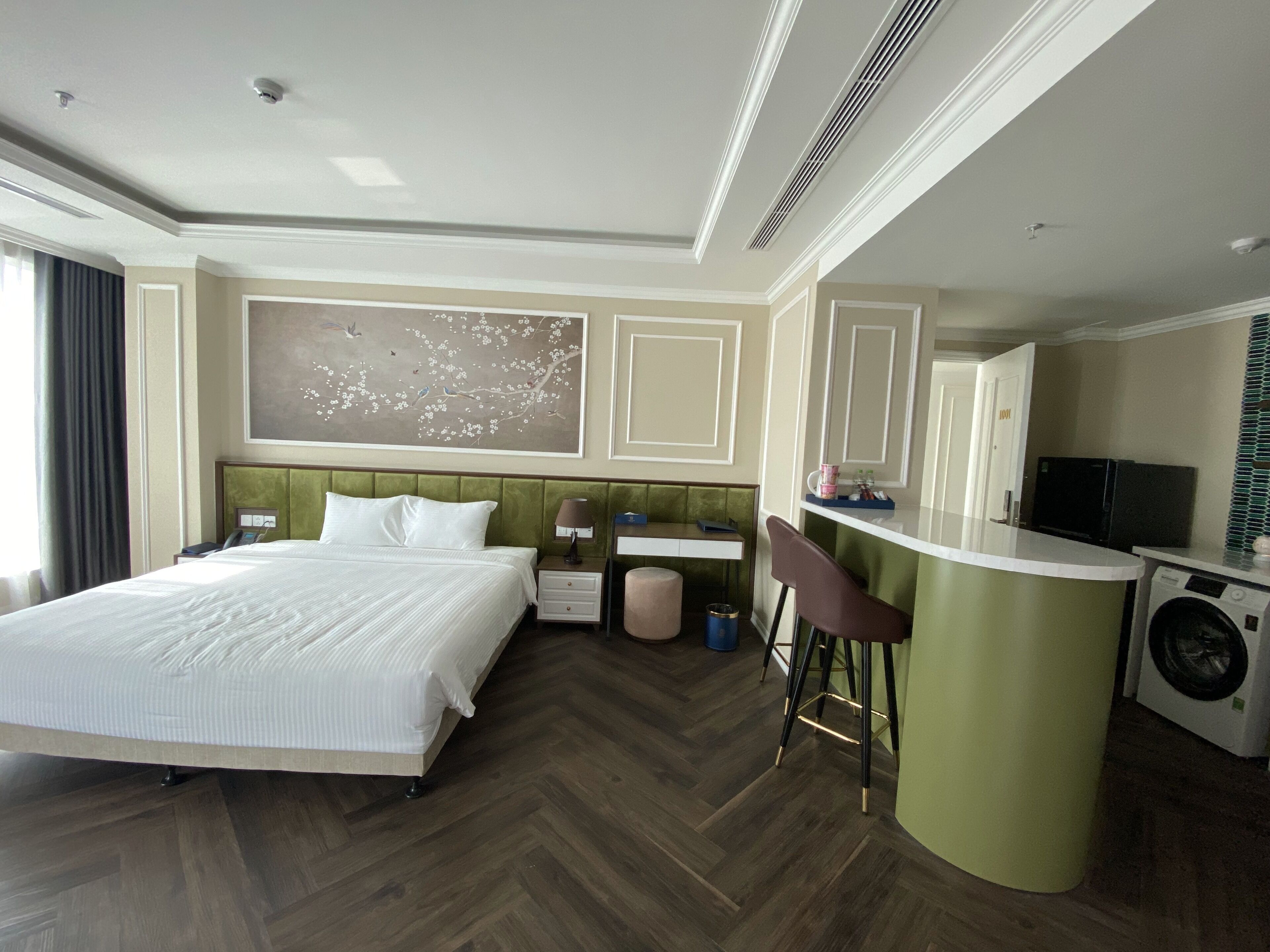 A hotel room with a large bed, a desk, a chair, a washing machine, and a painting on the wall.