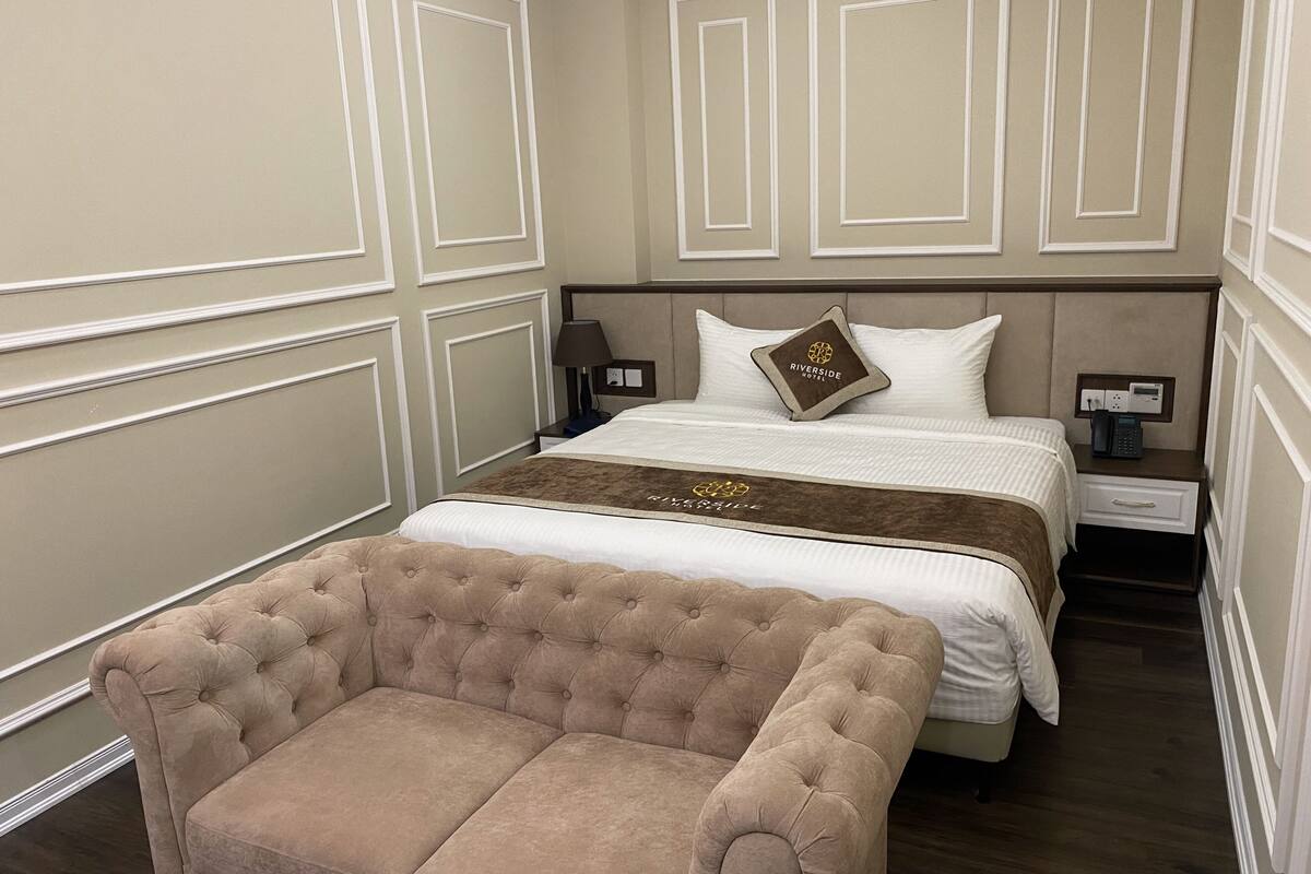 Una habitación de hotel con una cama grande, un sillón capitonado, una mesita de noche con una lámpara y otro atril con un teléfono.