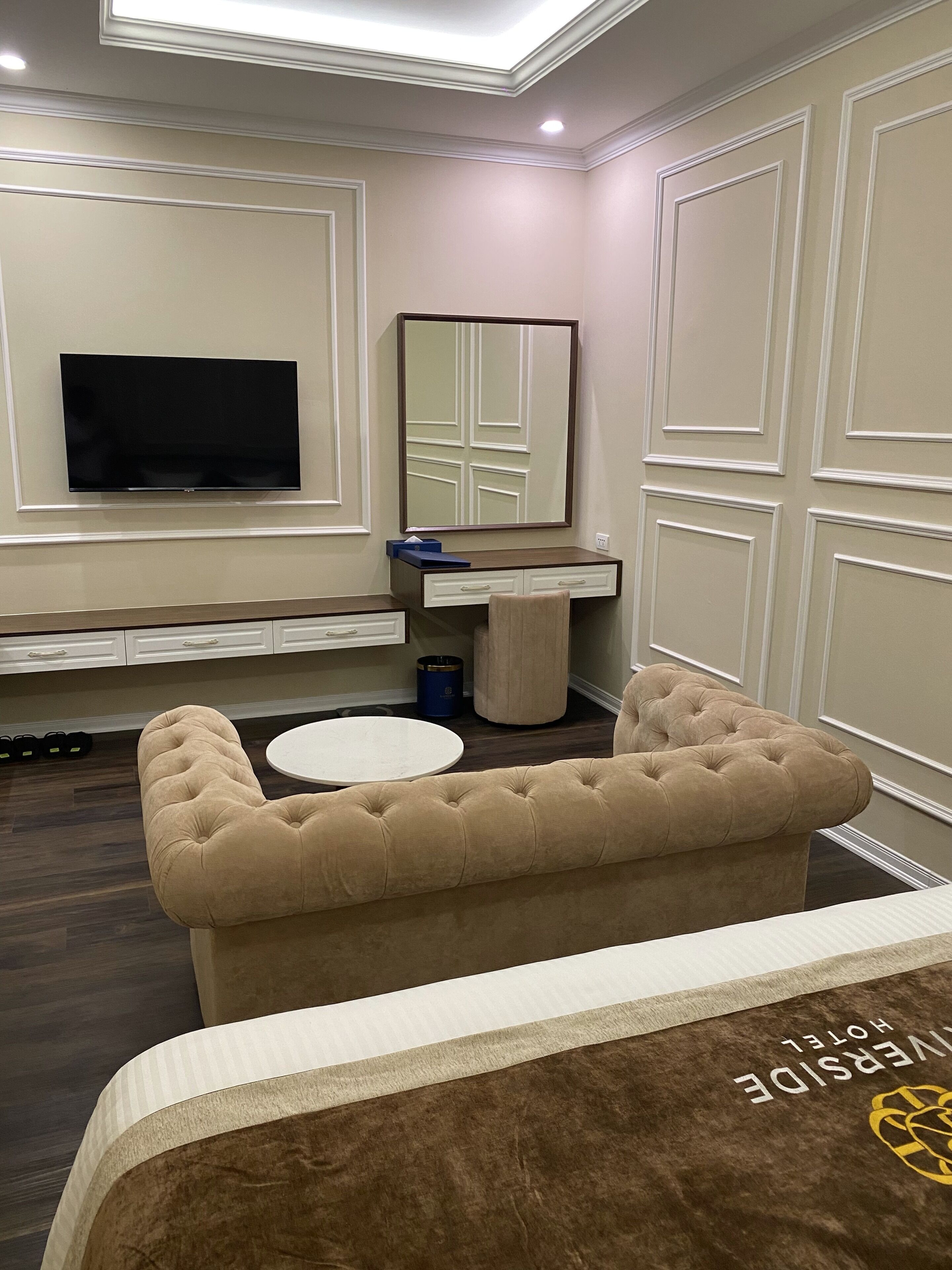 A hotel room with a large flat-screen TV mounted on the wall, a round white table, a tufted beige sofa, and a bed with a brown headboard.
