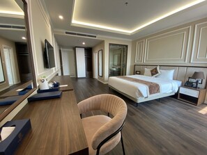 A modern hotel room with a large bed, a desk with a chair, a bedside table with a lamp, and a large mirror.