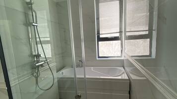 A modern bathroom with a glass-enclosed shower, a bathtub, and a window with blinds.
