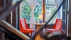 A room with two red upholstered chairs, a wooden table, a lamp, and a view of greenery through large windows.